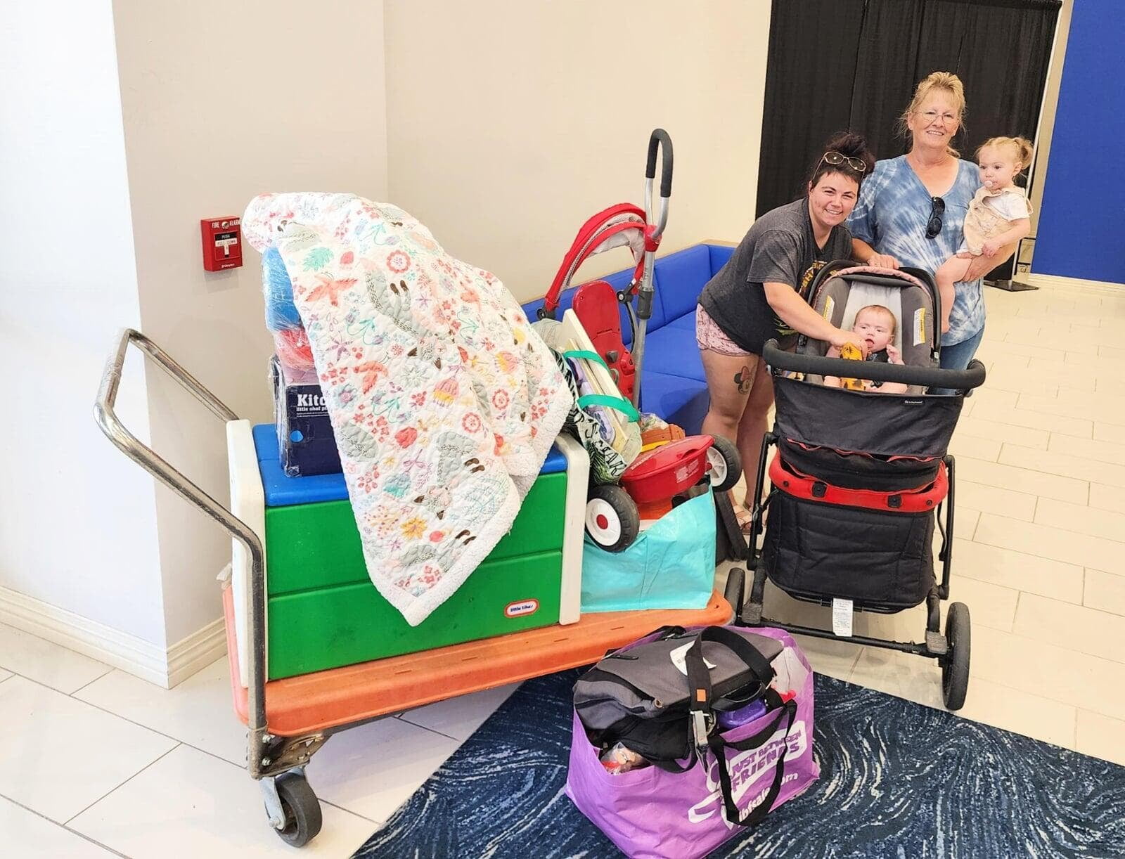 2 smiling women stand around a stroller with a baby in a carset while one holds a toddler. They are surrounded by items they purchased at the sale.