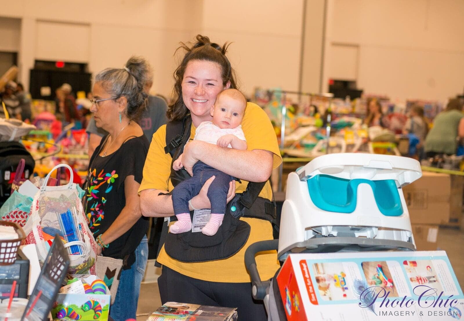 A mom with a big smile is holding her young child with shoppers in the background